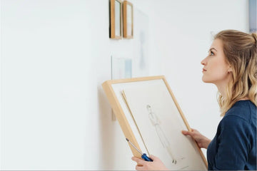 Frau hängt gerahmtes Line-Art Poster in Holzrahmen an weißer Wand auf - praktische Anleitung zum richtigen Positionieren und Aufhängen von Wandbildern